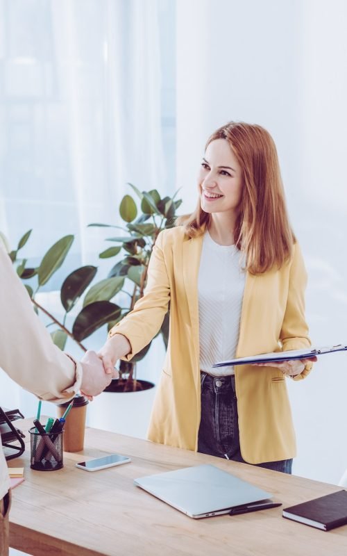 happy-recruiter-holding-clipboard-and-shaking-hands-with-employee-in-modern-office.jpg