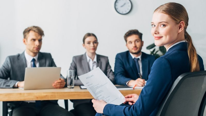 selective-focus-of-employee-looking-at-camera-while-holding-resume-near-recruiters.jpg